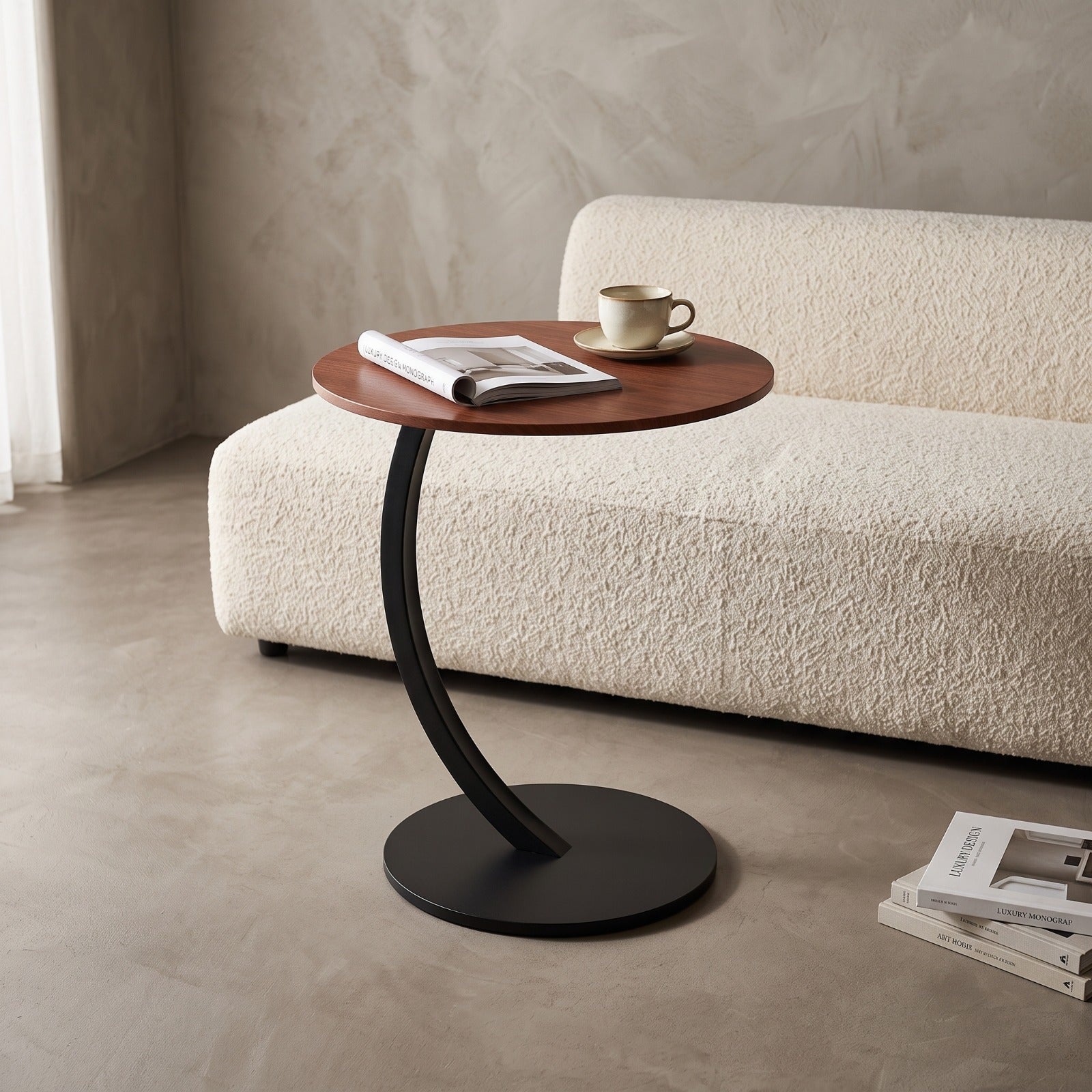 A modern round side table with a warm brown wood-grain top and a curved black metal base, styled in a minimalist living room next to a textured cream boucle sofa.