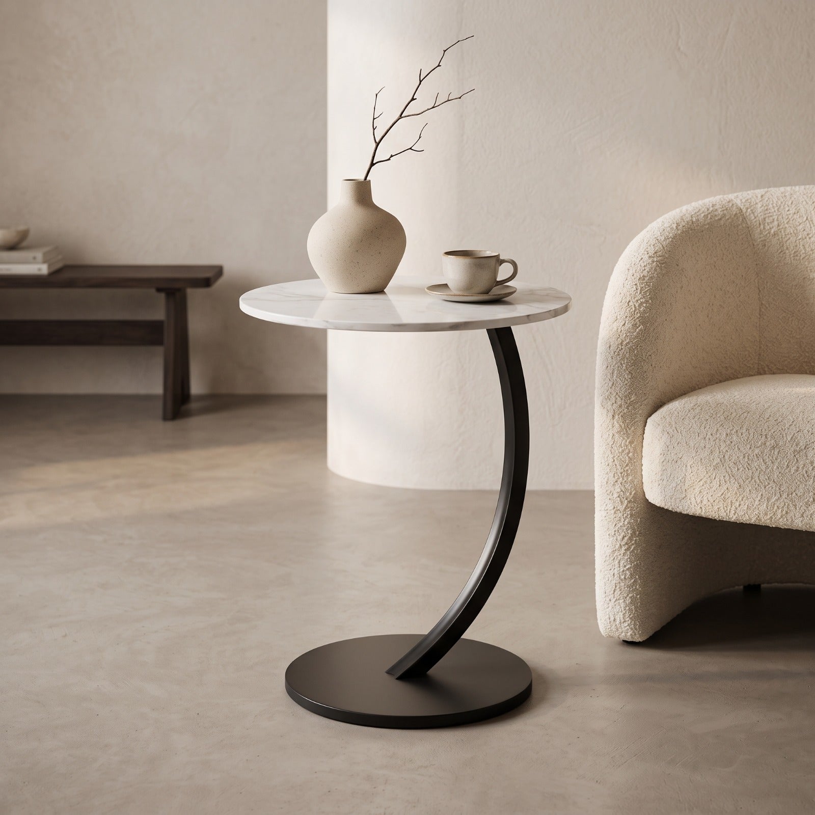 A modern round side table with a white marble top and a curved black metal pedestal base, styled in a minimalist room with a beige armchair, a ceramic vase, and a cup on a saucer.