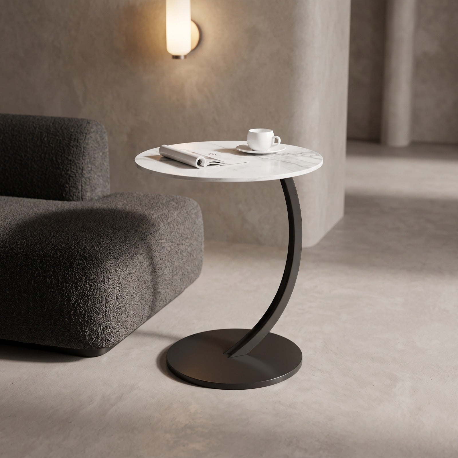 A modern round side table with a white marble top and a dark curved metal pedestal base, styled in a minimalist room next to a dark gray textured sofa.