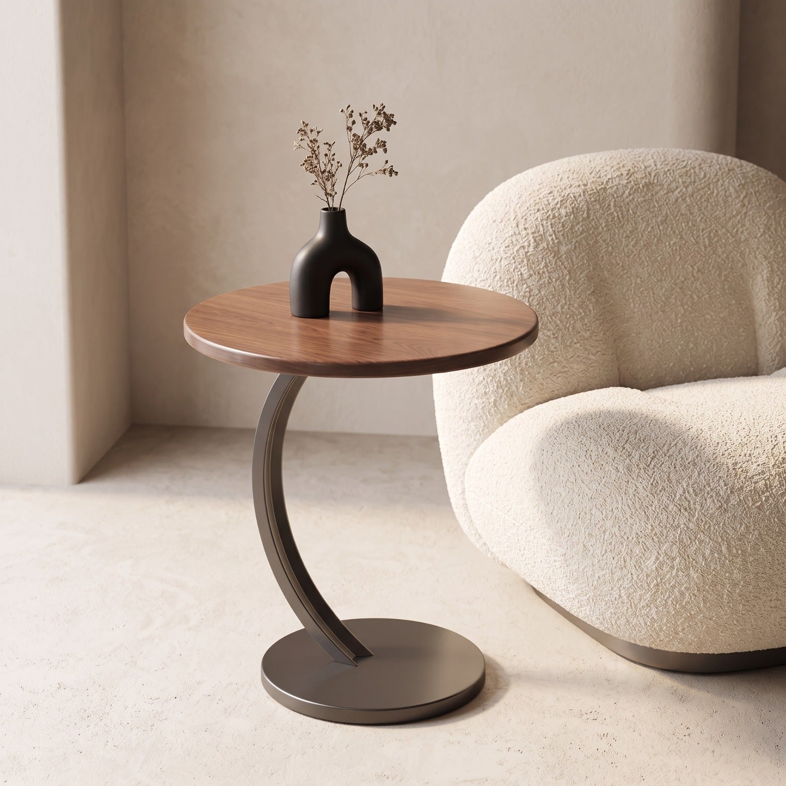 A modern round side table with a medium brown wood top and a curved dark metal base, styled next to a cream boucle armchair with a small black vase on top.