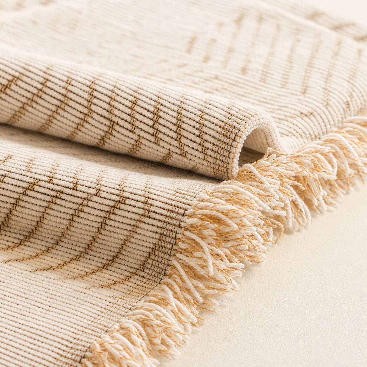 A close-up of a woven throw blanket in cream and tan with a fringed edge, folded to show texture and pattern.