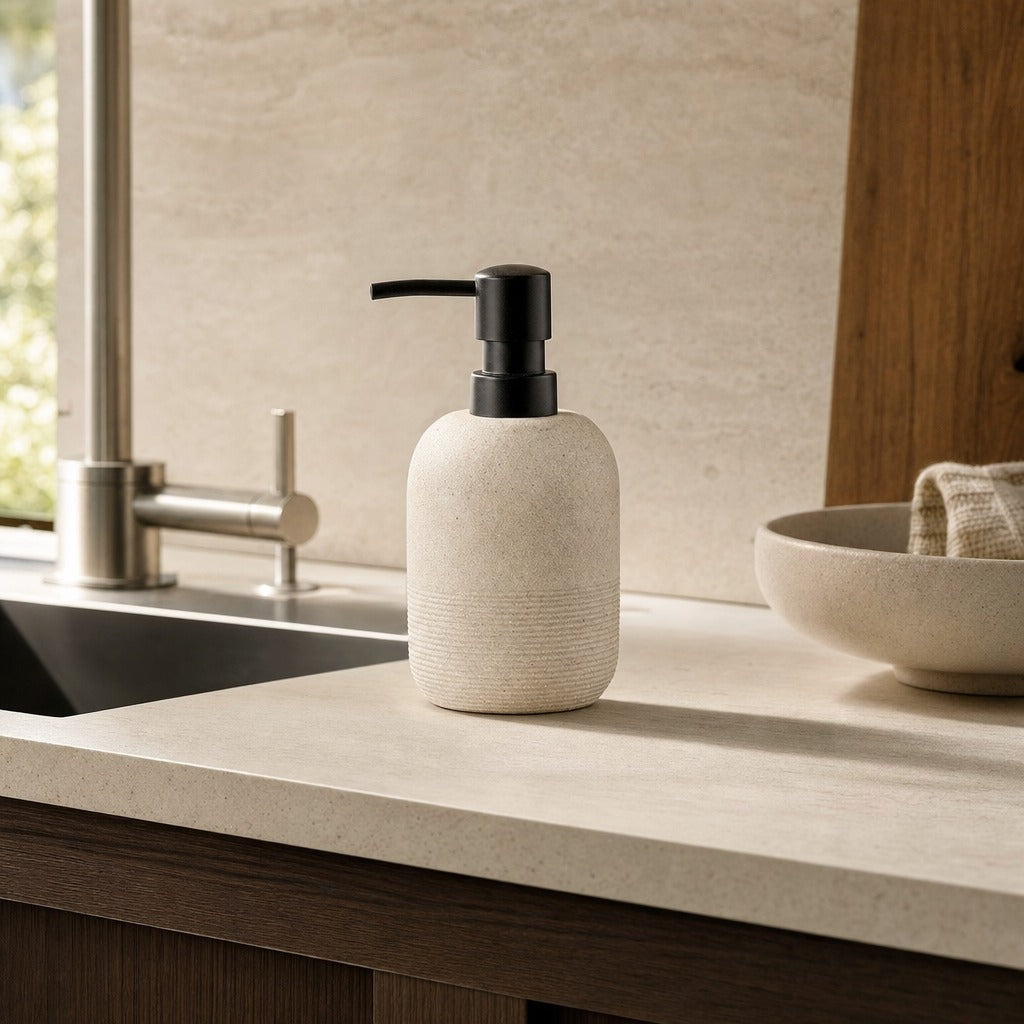 A textured cream soap dispenser with a matte black pump bottle, set on a light beige countertop next to a sink and wooden cabinetry.
