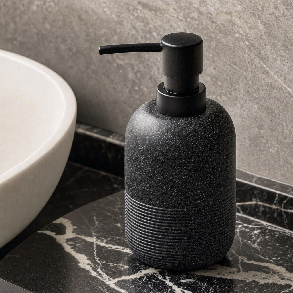 A matte black textured soap dispenser with a ribbed lower section and a black pump, shown on a dark marble countertop next to a white sink against a gray stone wall.