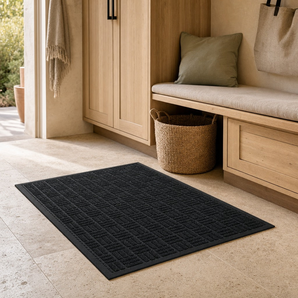 A dark charcoal gray textured rectangular mat with a raised geometric pattern, shown on a light beige stone floor in a modern entryway setting next to a wooden bench and storage cabinet.