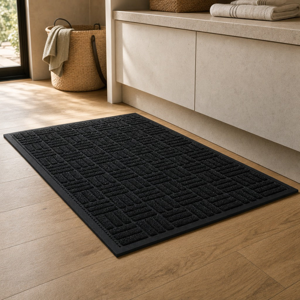 A black rectangular bath mat with a raised geometric pattern, shown on a light wood floor in a bathroom setting next to a beige vanity.