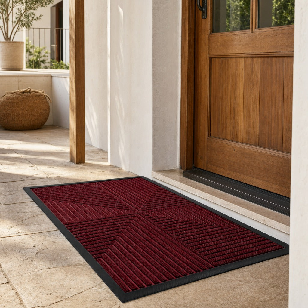 A rectangular entryway doormat in deep burgundy with a black border, shown on a stone patio in front of a wooden front door.
