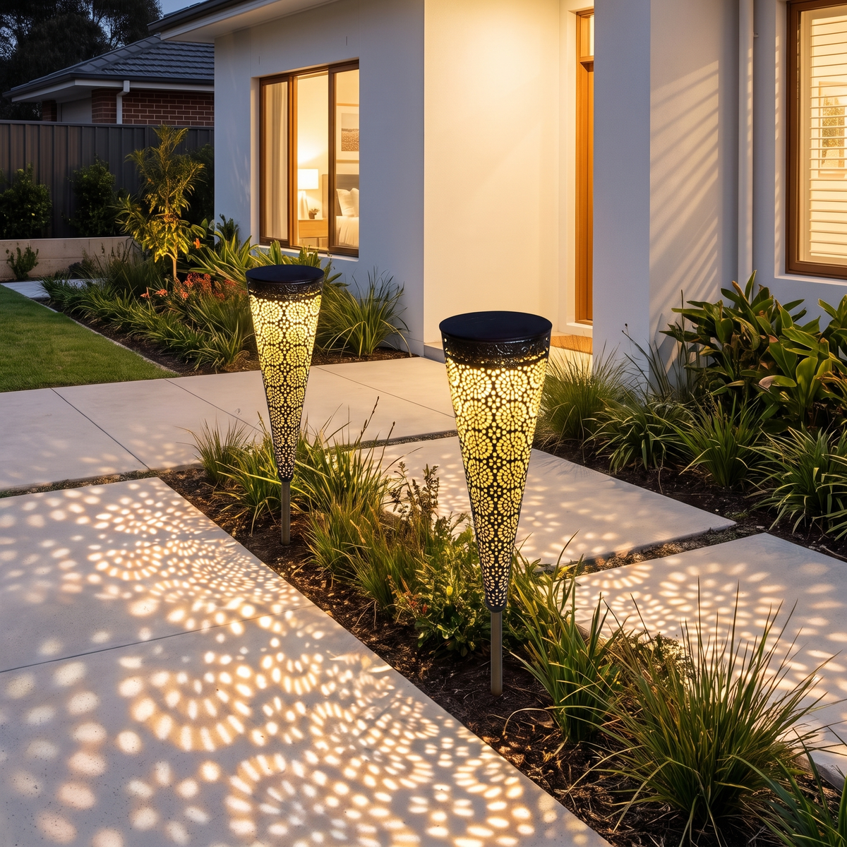 Two Bili Solar Garden Lights casting lace light patterns on grass by a concrete path of a white rendered home at night