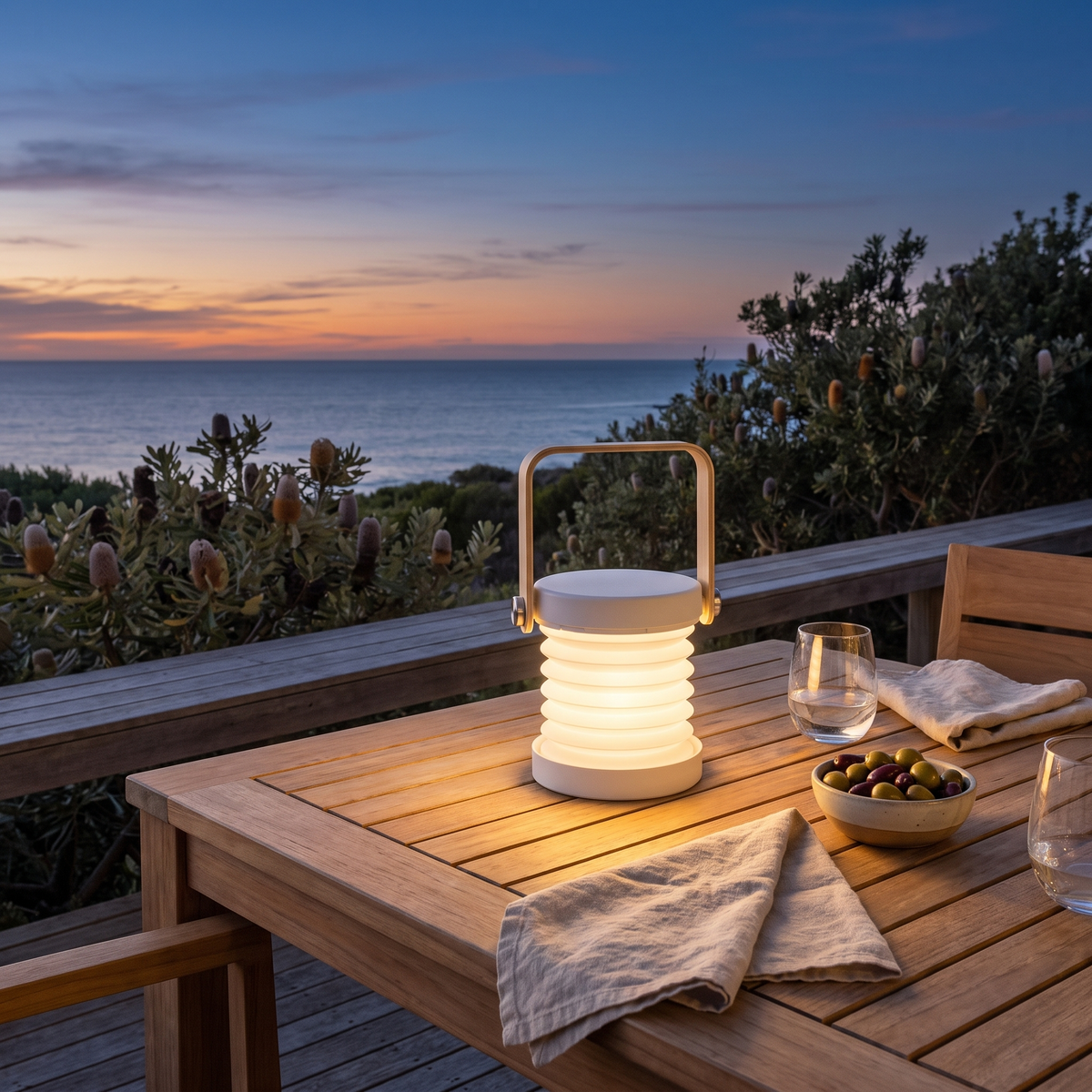 Brae folding travel lantern in white glowing on an outdoor dining table on a coastal deck at sunset