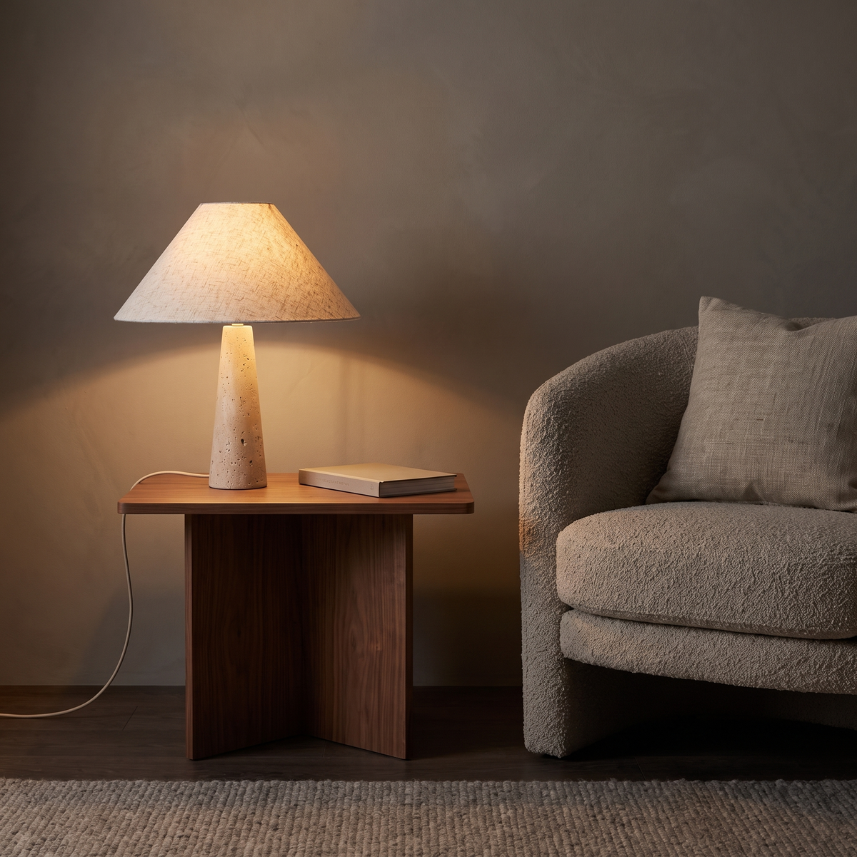 Fingal travertine table lamp lit on a walnut side table in a reading corner at night