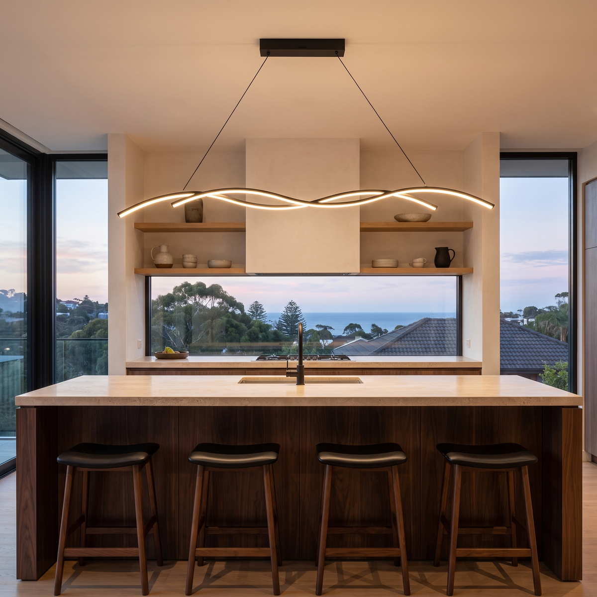 Grajagan black 150 cm wave pendant over a walnut kitchen island at dusk with a hillside sea view