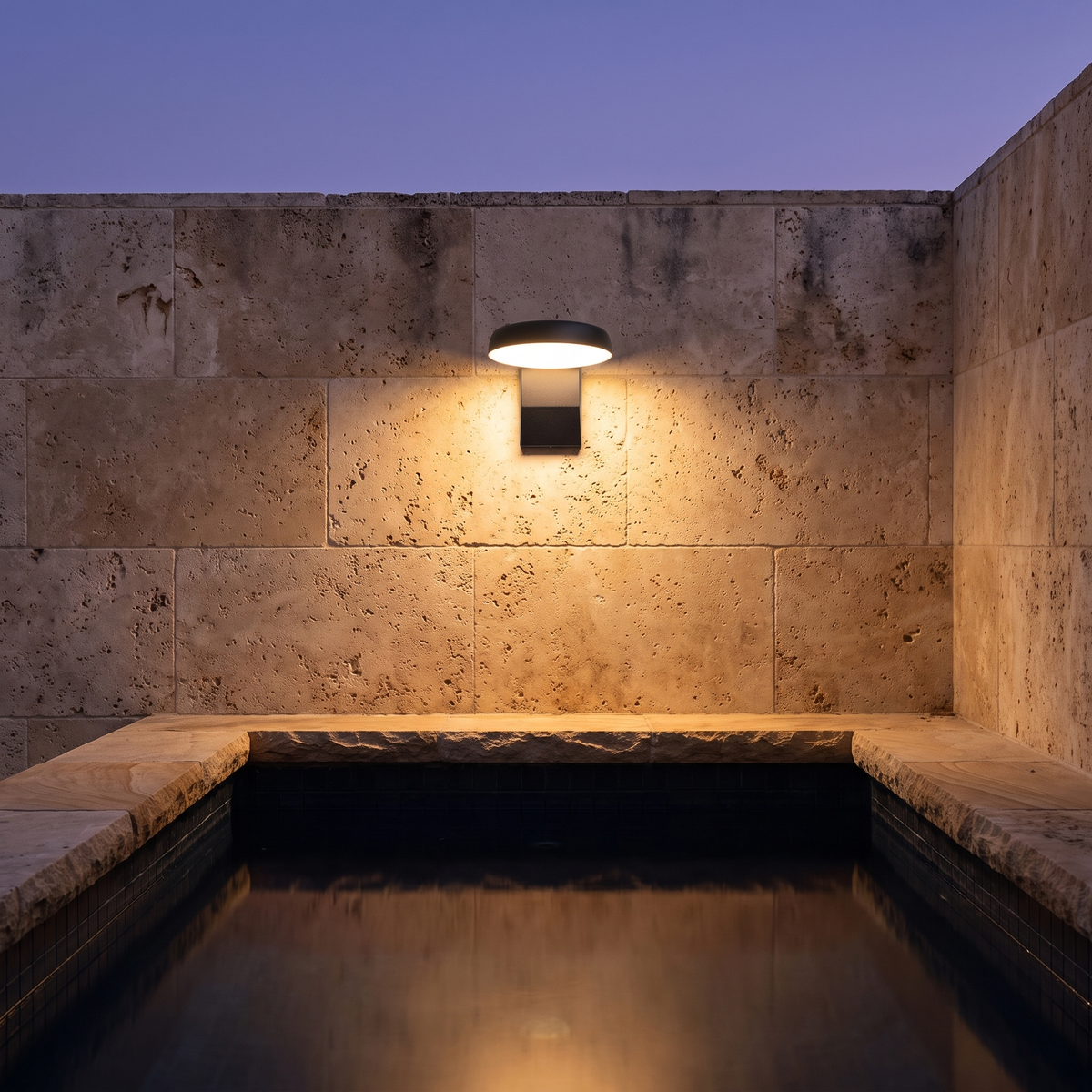 Kala Modern Outdoor Wall Light round black on travertine courtyard wall by a plunge pool, warm white light
