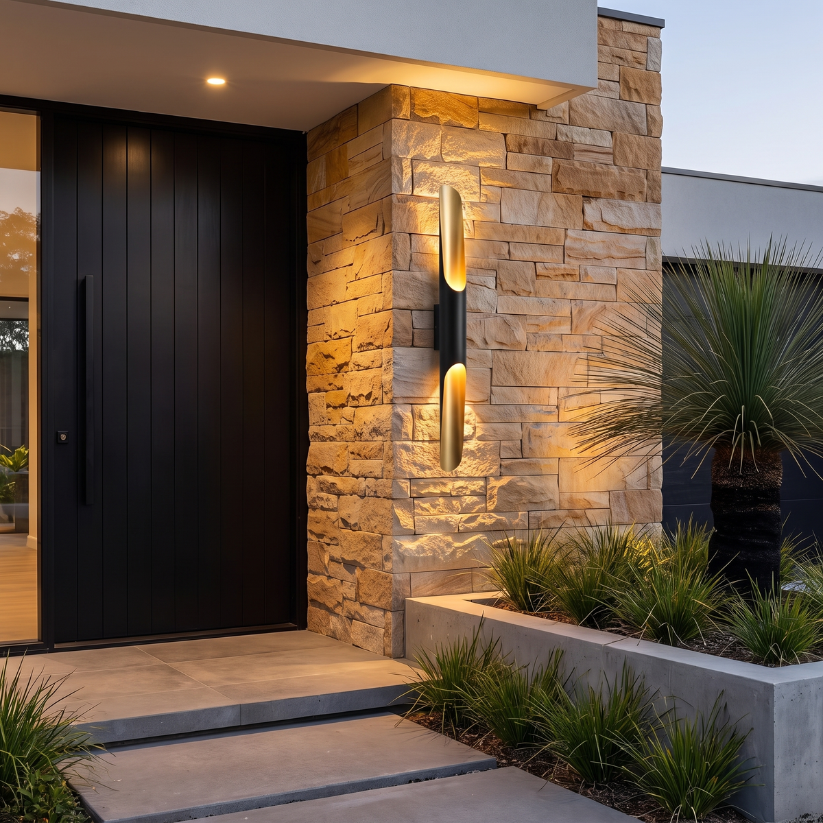 Jarra black and gold outdoor wall light, single tube, on stacked sandstone beside a pivot door at dusk