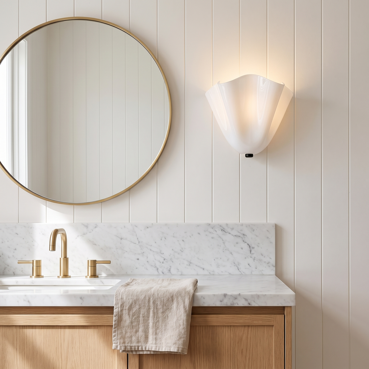 Kira artistic wall sconce in white glass lit beside a round mirror in a marble coastal bathroom