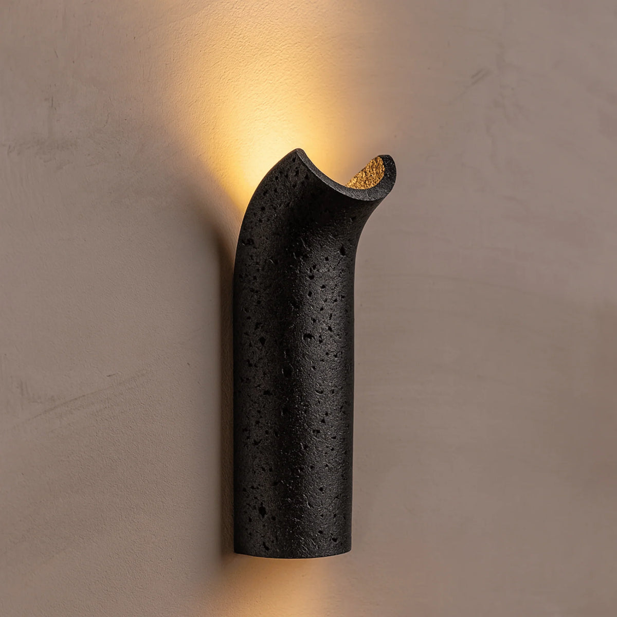 Pasa travertine wall lamp lit on a plaster wall, showing the dark stone and scoop top