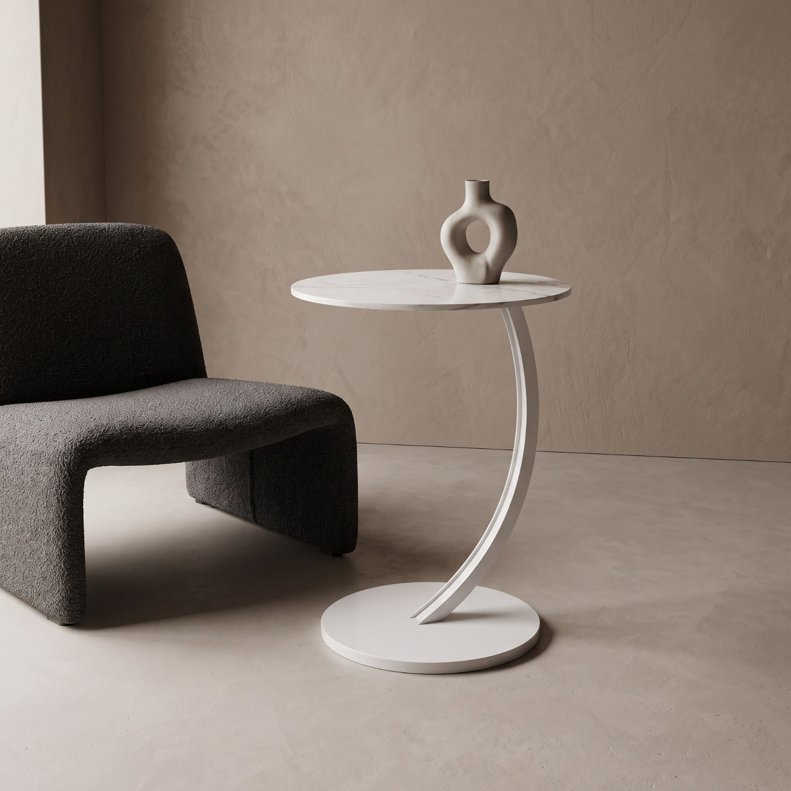 A modern white side table with a curved pedestal base and a small abstract ceramic vase on top, shown beside a dark gray upholstered chair in a minimalist beige room.