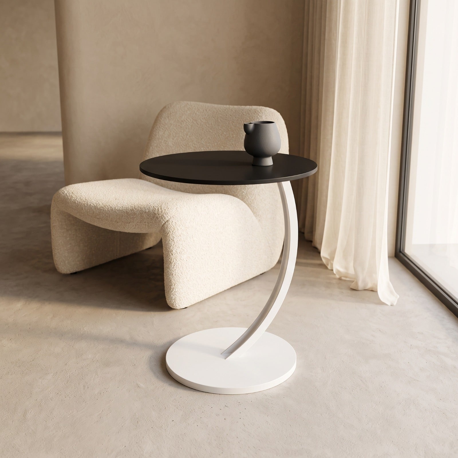 A modern round side table with a black top and a curved white pedestal base, styled in a minimalist beige room next to a cream bouclé chair and a small dark gray ceramic pot.
