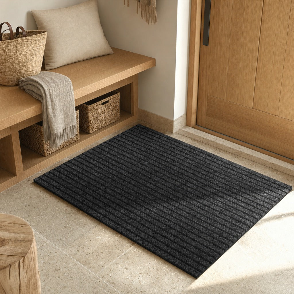 A dark charcoal gray ribbed entryway mat placed on a light stone floor in front of a wooden door, with a wooden bench, woven baskets, and a throw pillow visible in the room setting.