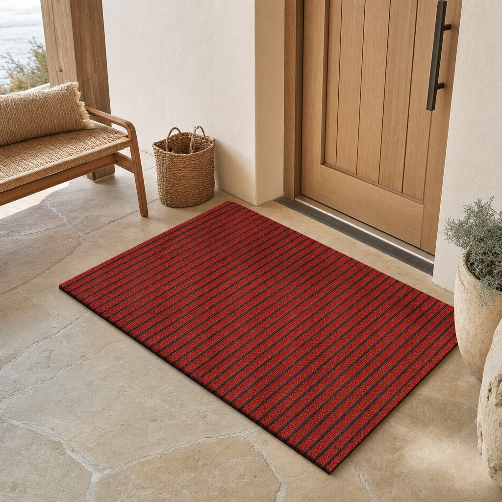 A rectangular red entryway mat with thin dark vertical stripes lies on a light stone floor in front of a wooden door, with a woven bench, basket, and potted plant nearby.
