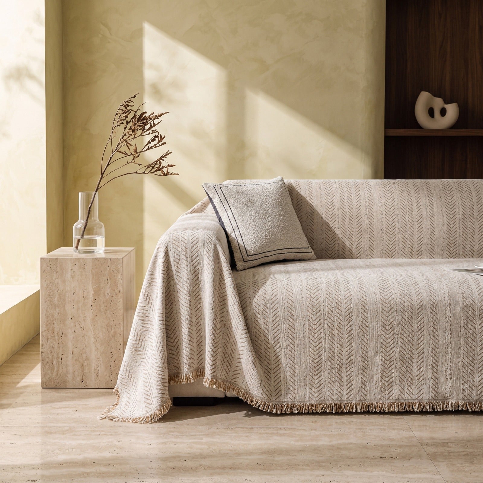 A beige textured throw blanket with a chevron pattern and fringe is draped over a sofa in a sunlit living room. A matching pillow rests on the sofa, next to a stone side table holding a glass vase with dried branches.