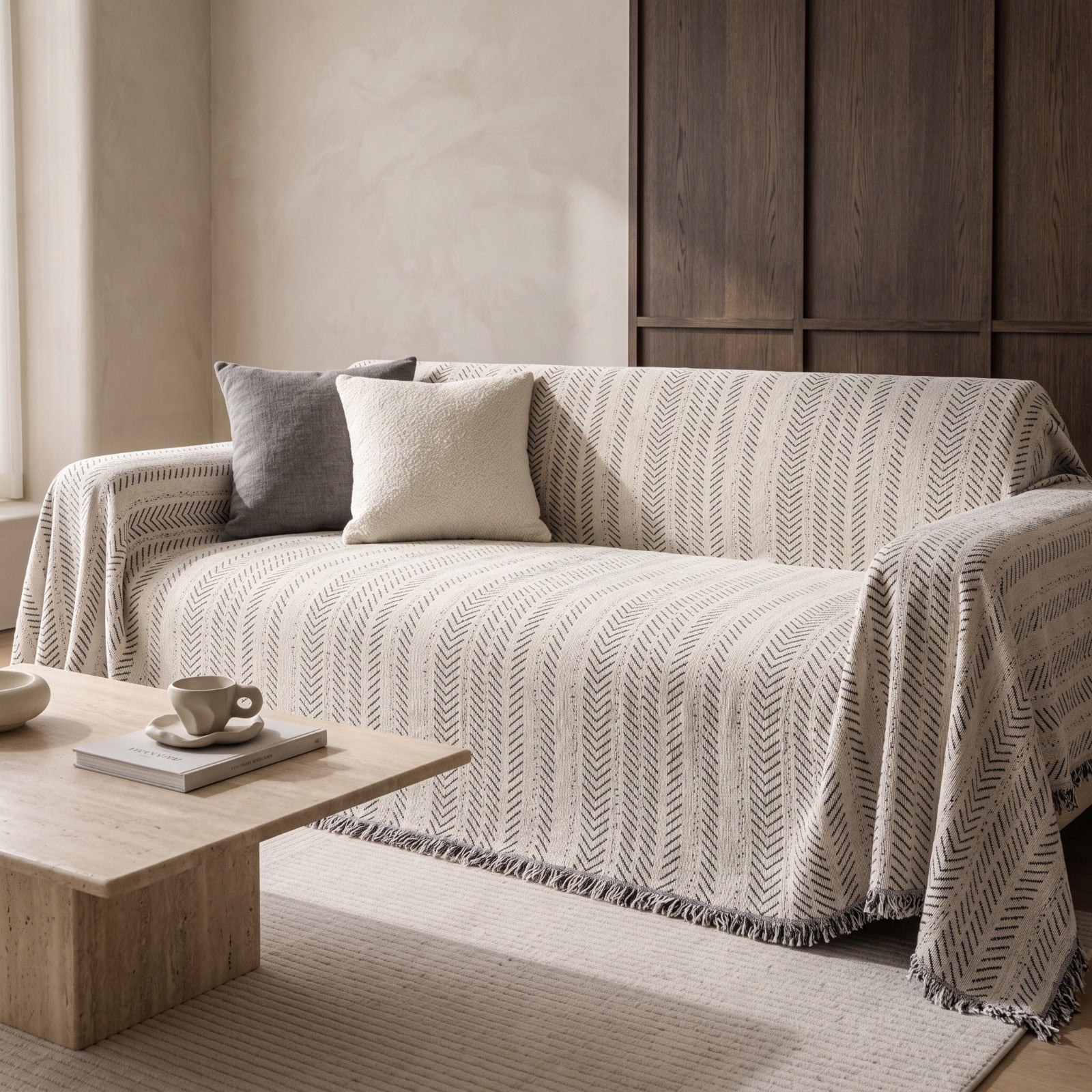 A beige and gray chevron-patterned throw blanket with fringe trim draped over a sofa in a modern, minimalist living room setting.