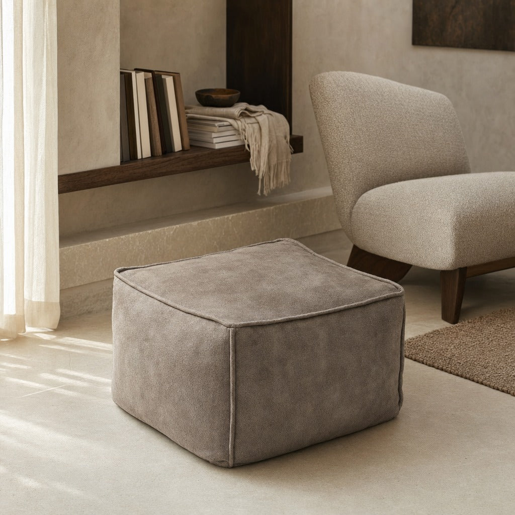 A square upholstered ottoman in a warm taupe-gray fabric, styled in a modern living room setting next to a matching armchair.