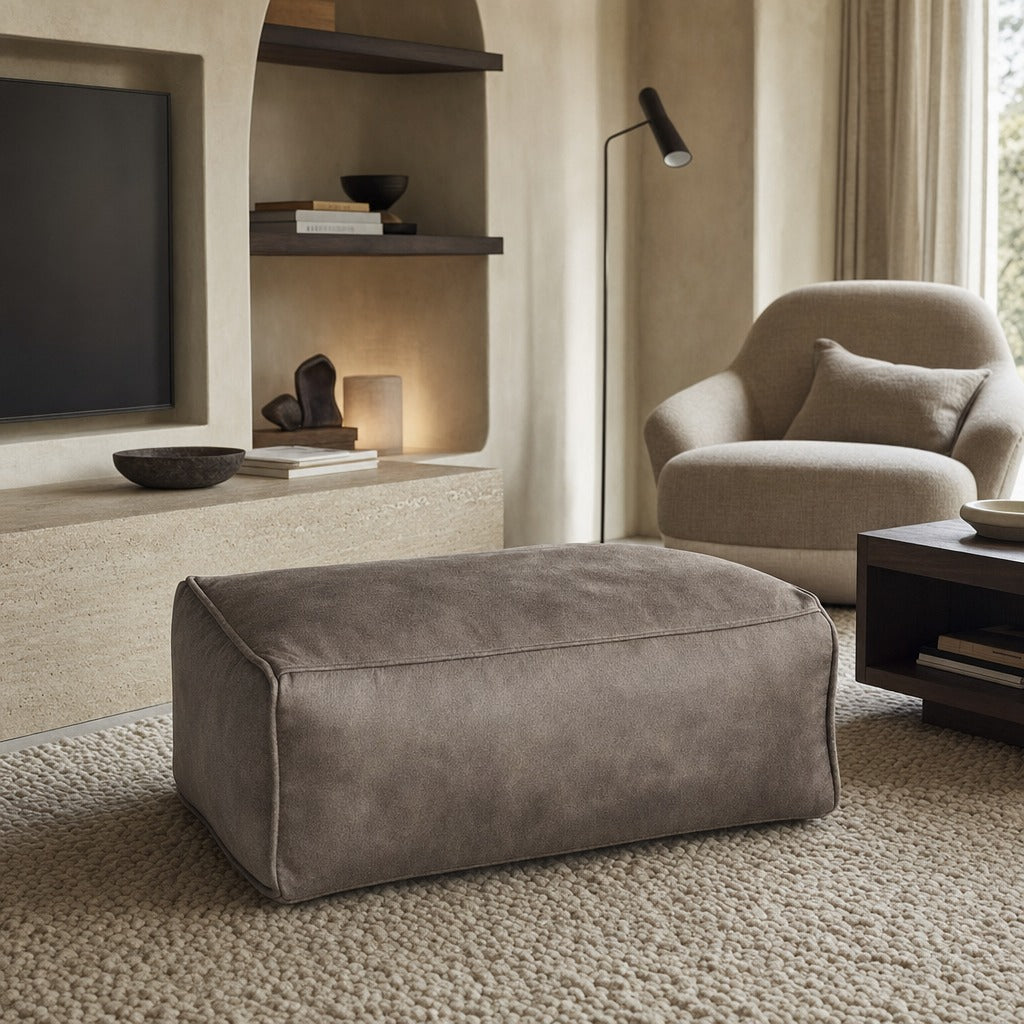 A rectangular upholstered ottoman in a warm taupe-brown suede-like fabric, placed in a modern living room setting with a matching armchair, textured rug, and built-in shelving.