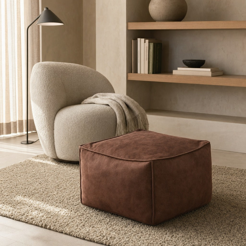 A square brown suede or microfiber ottoman in the foreground of a styled living room scene, with a cream bouclé armchair, floor lamp, books, and shelving in the background.