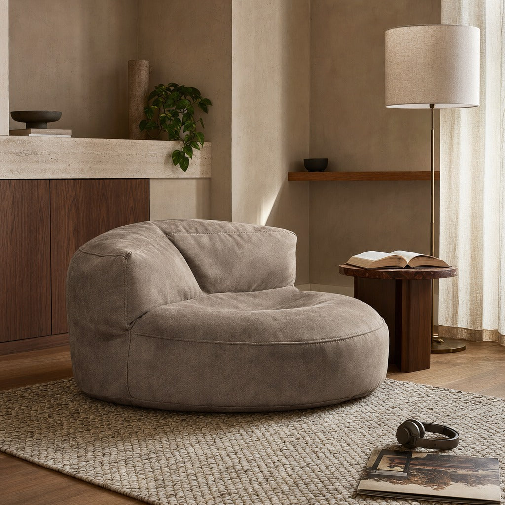 A round, low-profile lounge chair in a taupe bouclé fabric, styled in a warm, modern minimalist living room setting with a wooden side table, floor lamp, and textured rug.