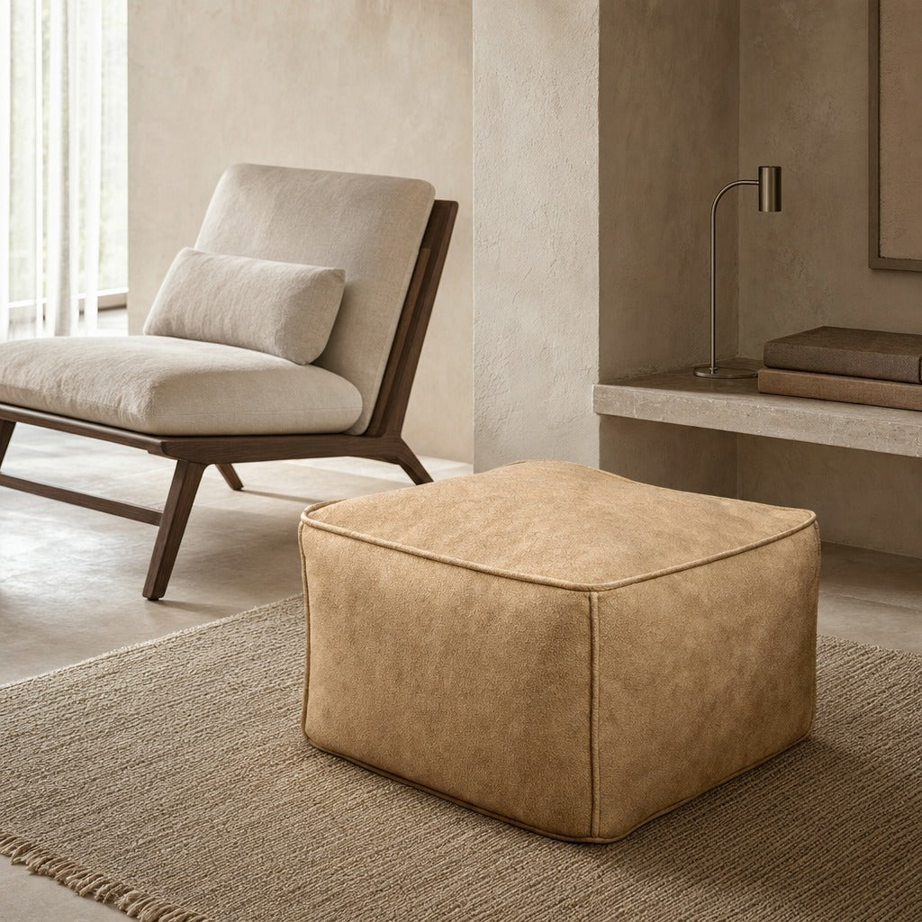 A square tan upholstered ottoman in a modern, minimalist room setting with a beige armchair and textured rug.