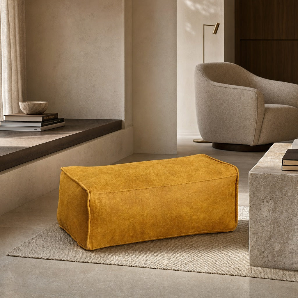 A mustard yellow velvet ottoman in a modern, minimalist living room setting with a textured beige armchair, concrete tables, and a neutral rug.
