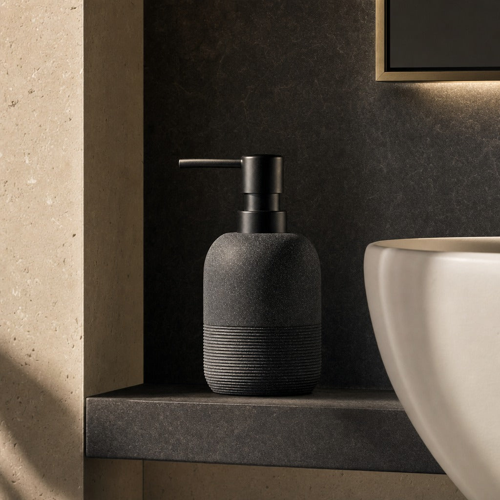 A dark charcoal gray textured soap dispenser with a matte black pump, resting on a dark gray bathroom ledge next to a white ceramic sink.