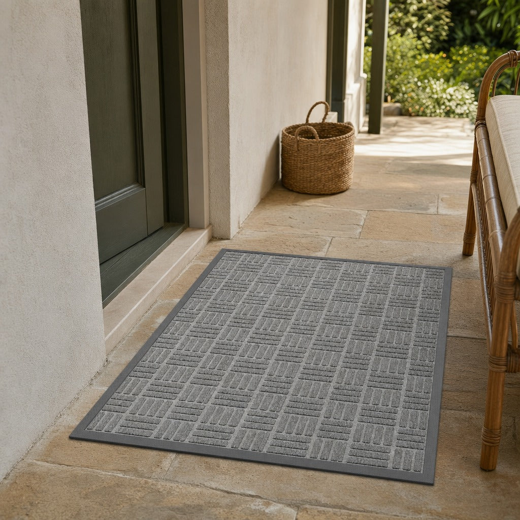 A gray rectangular outdoor doormat with a textured basketweave pattern, shown on a stone patio at the entrance to a home next to a door and a woven basket.