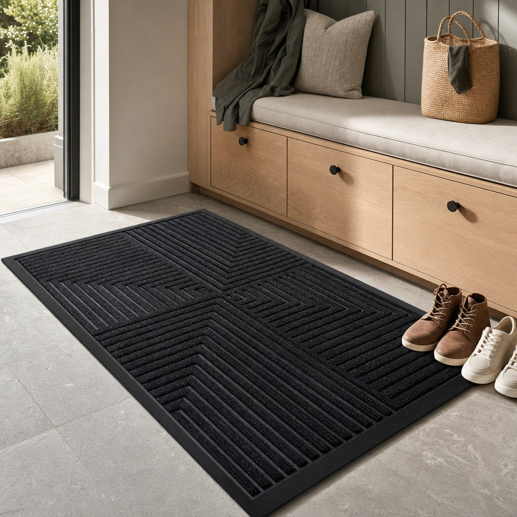 A black rectangular entryway rug with a raised geometric pattern, shown on a light gray tile floor in a modern entryway with a wooden bench, shoes, and a bag nearby.