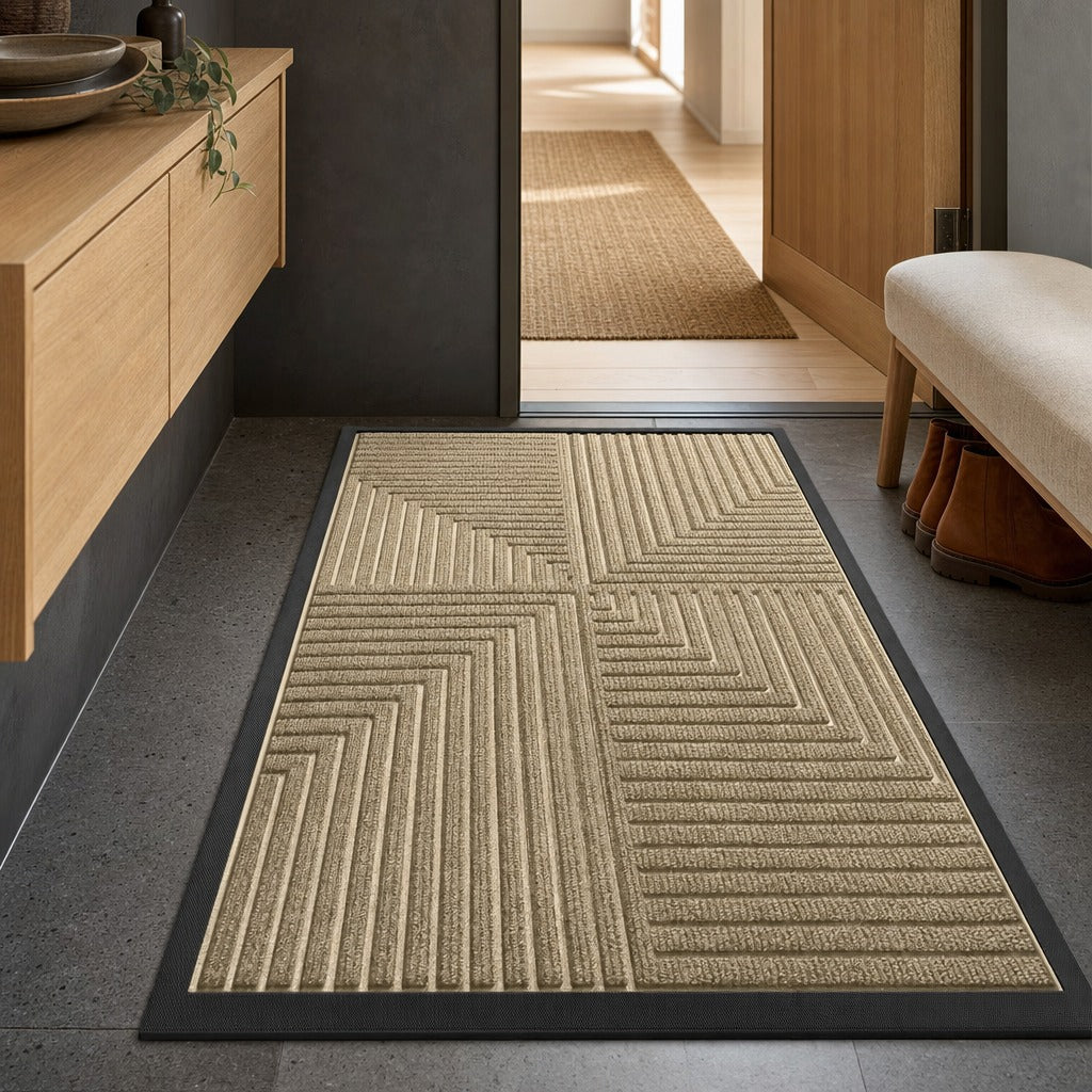 A rectangular entryway rug with a textured geometric chevron pattern in beige and natural fiber tones, bordered in black, shown on a gray floor in a modern interior setting.