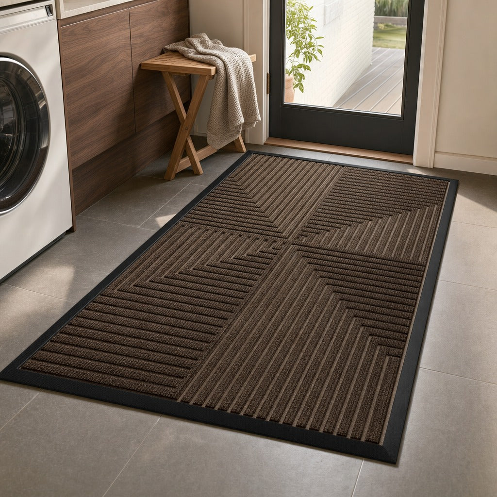 A rectangular brown textured doormat with a black border and geometric ridged pattern, shown on a tiled floor next to a washing machine and doorway.
