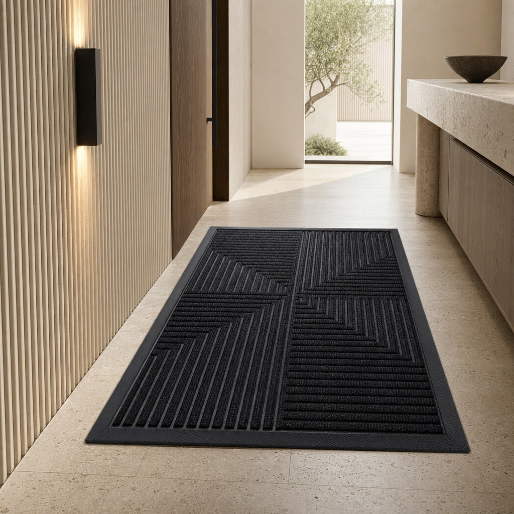 A rectangular black entryway mat with a textured geometric chevron pattern, shown on a light beige stone floor in a modern hallway.