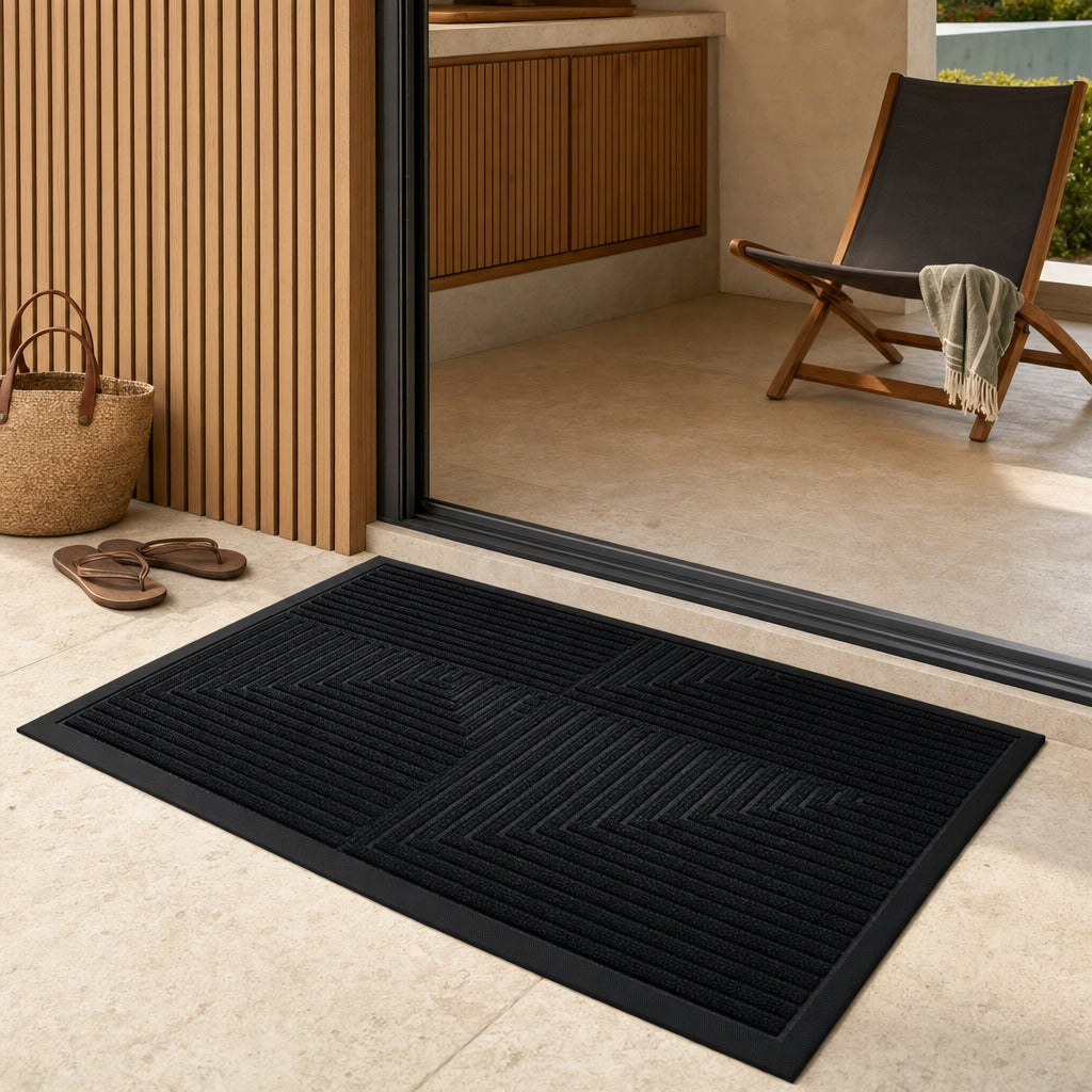 A black rectangular doormat with a raised geometric chevron pattern, shown on a light beige tiled patio floor in front of a glass sliding door. A woven basket, sandals, and a wooden lounge chair are visible in the background.