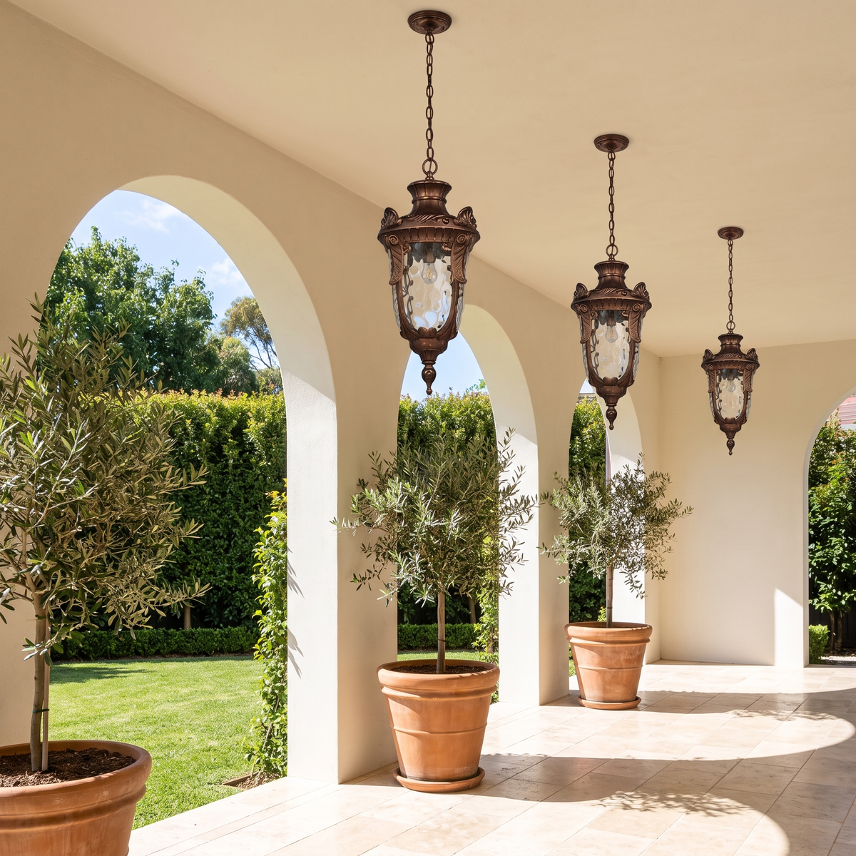 Three Anita vintage lantern pendants in bronze hanging in an arched rendered loggia with potted olives