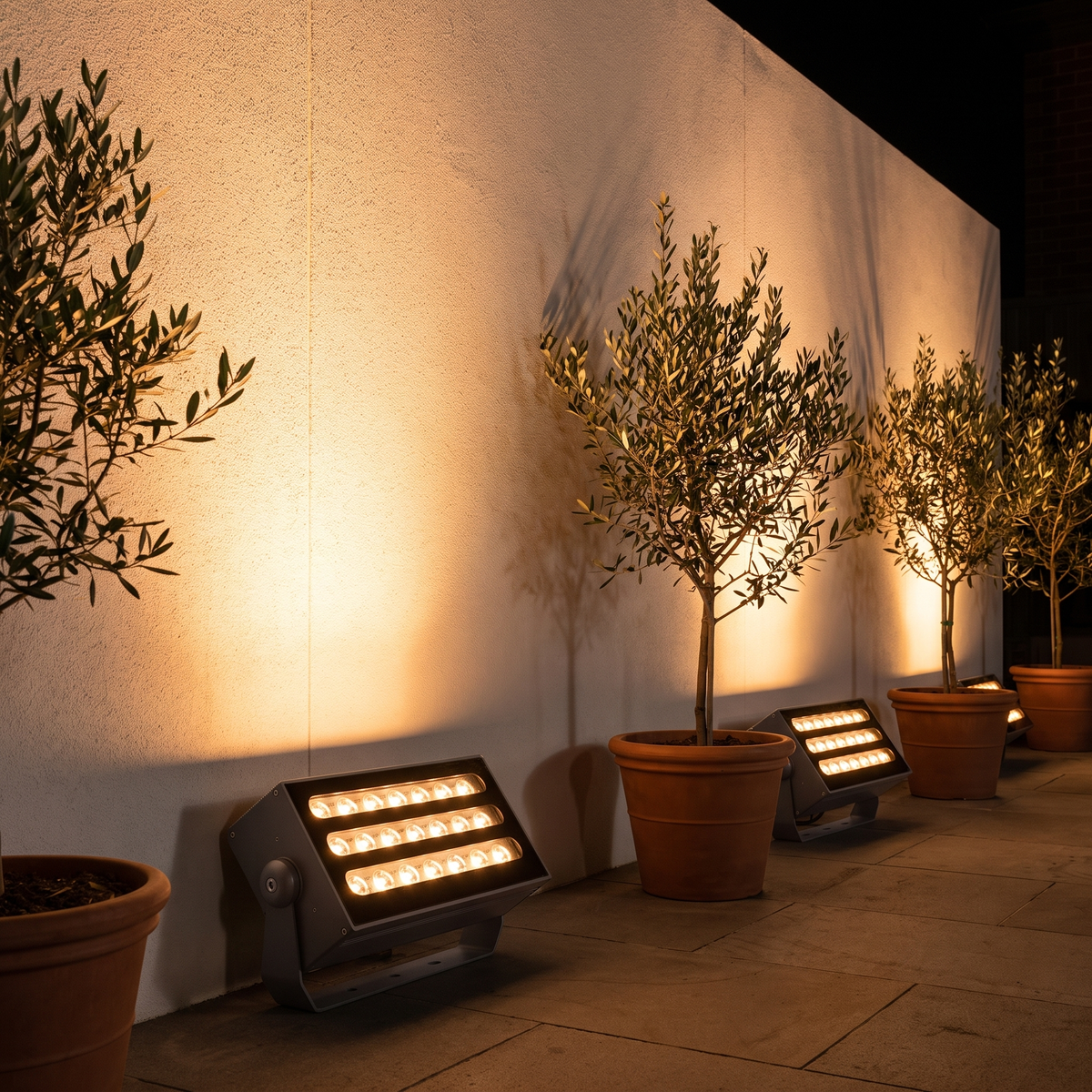 Two Honnibal RGB LED flood lights washing a rendered courtyard wall in amber with potted olive trees