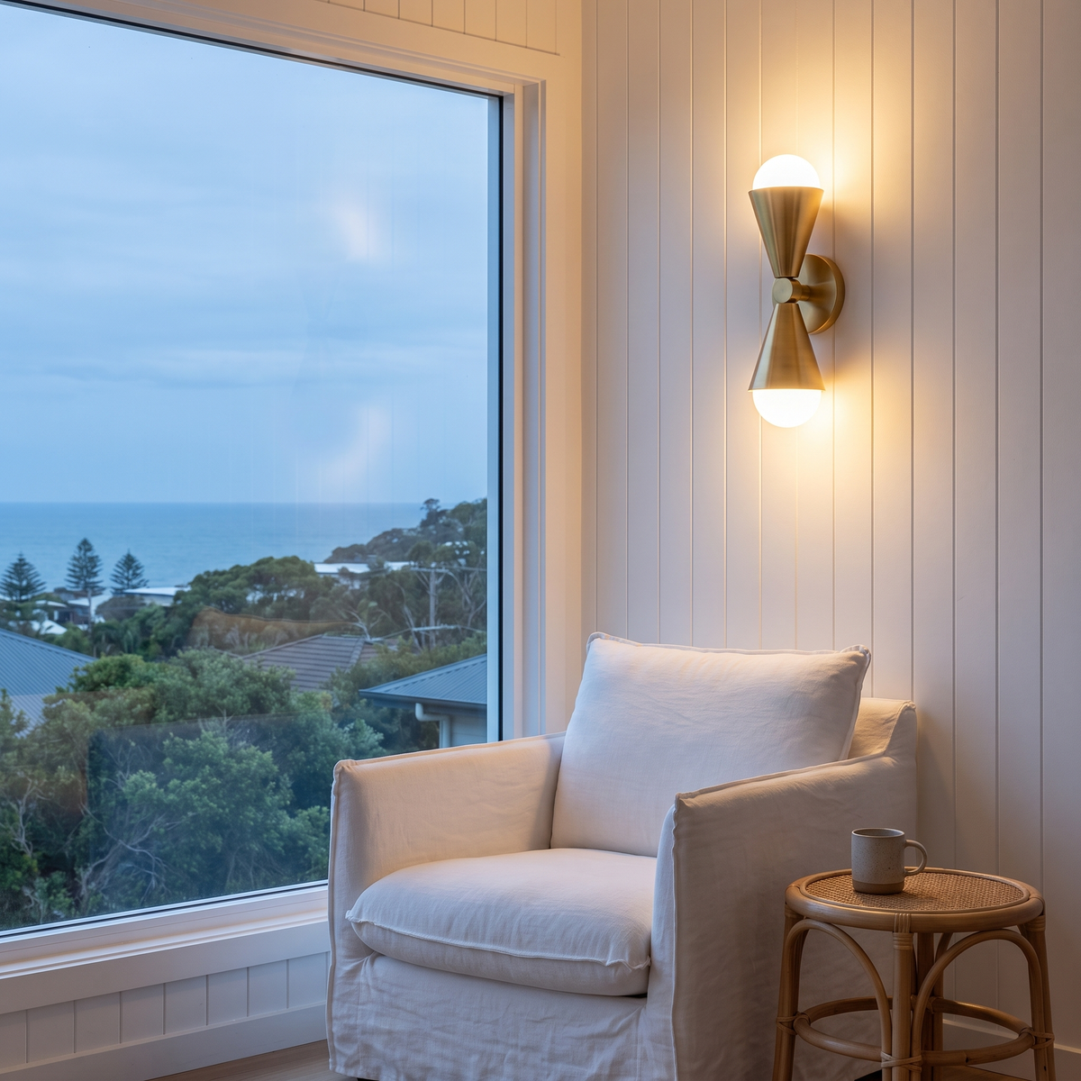 Lura twin wall sconce glowing above a linen armchair in a reading nook with an ocean view at dusk