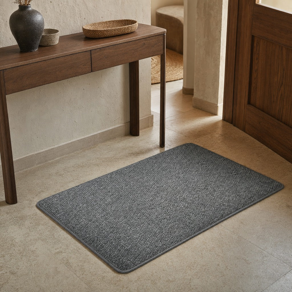 A rectangular gray textured entryway mat placed on a light beige tile floor in a styled interior setting with a wooden console table and door visible.